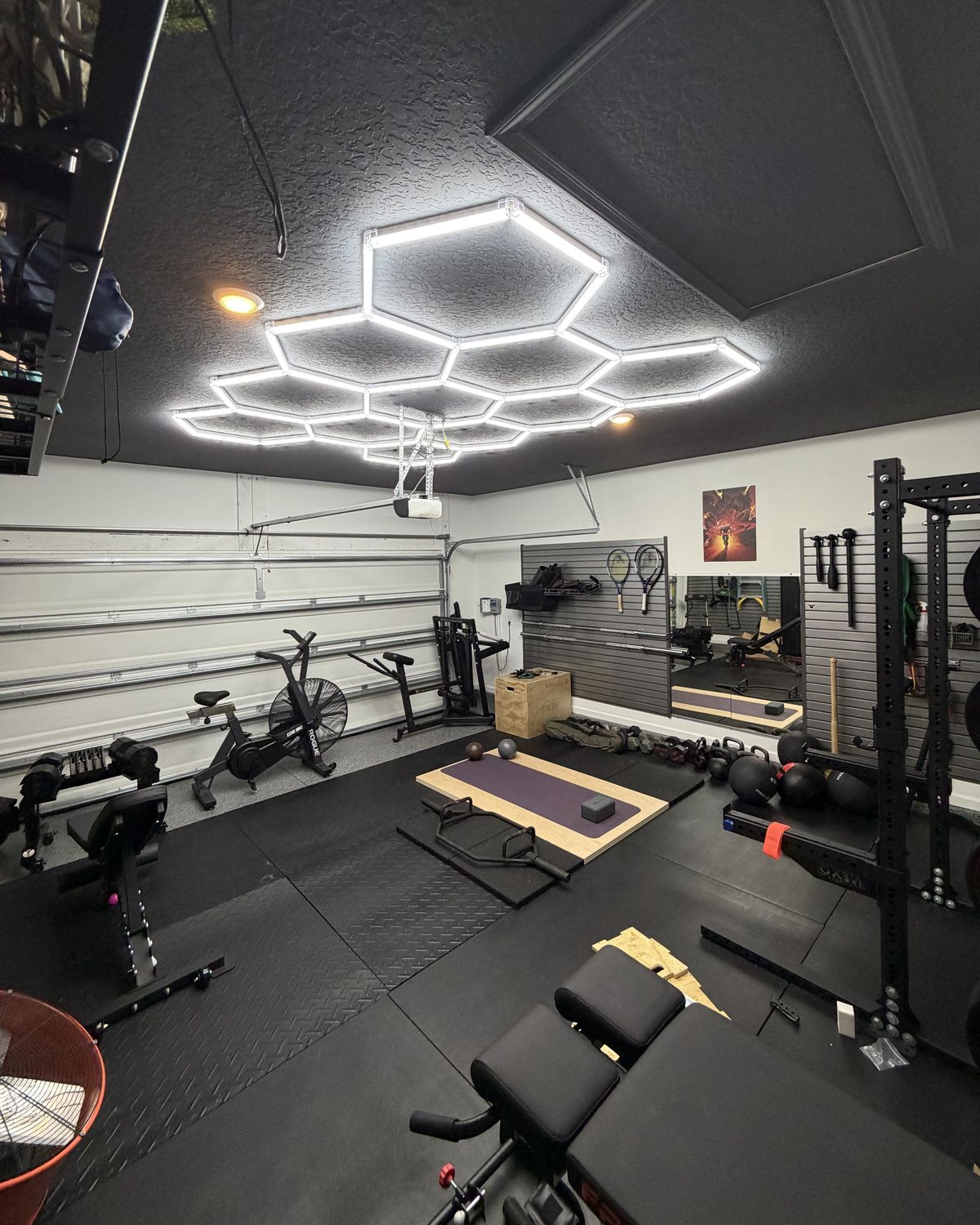A garage gym under hexagon lights, with a squat rack, lifting platform, air bike and plyo box